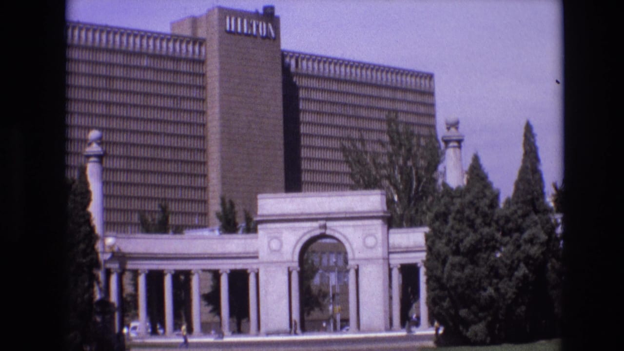 1974 Denver home-movie frame showing a tall Hilton hotel rising behind white archways, columns, street-level landscaping, and small pedestrians.