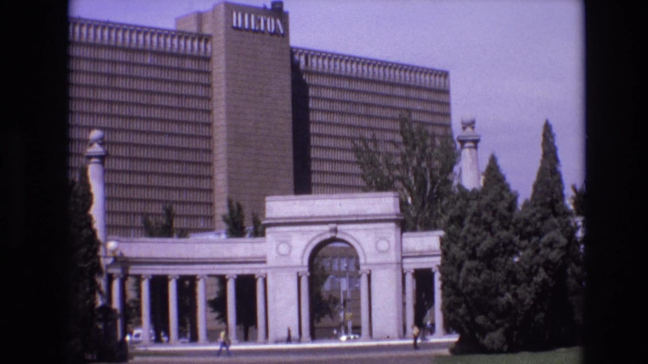 Hotel tower and entrance archway seen across a street with trees and pedestrians.
