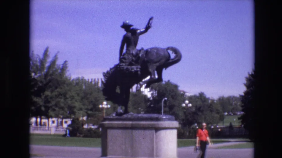 1974 Denver home-movie frame showing a bronze cowboy riding a bucking bronco statue on a stone pedestal, with lawn, trees, paths, and a pedestrian in a red shirt nearby.