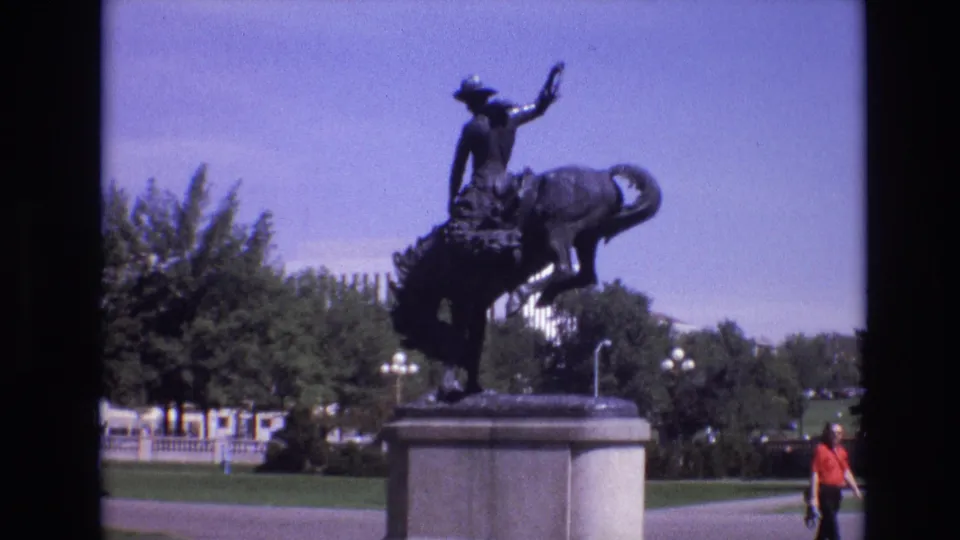 1974 Denver frame of the cowboy riding bronco statue on a pedestal, with landscaped grounds, trees, paved paths, and a pedestrian in a red shirt at right.