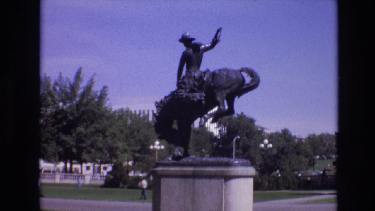 Bronze rider and bucking horse statue on a pedestal in a sunny park.