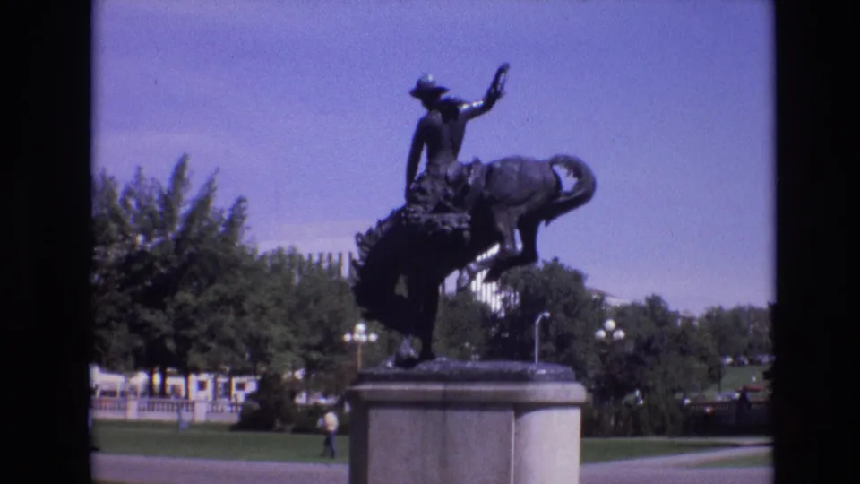 1974 Denver home-movie frame showing a bronze rider-and-horse statue on a stone pedestal with trees, lawn, lampposts, and distant pedestrians behind it.
