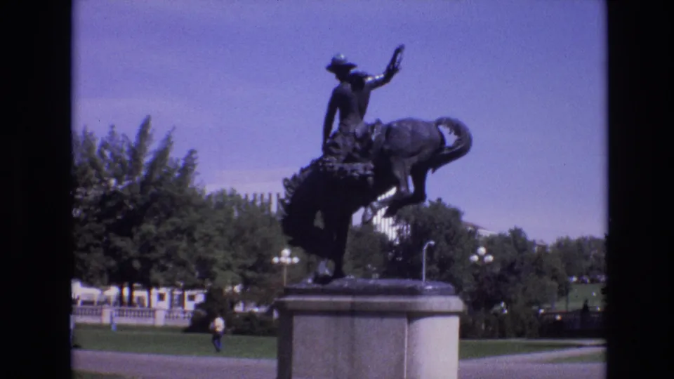 A 1974 Denver 8mm view holds on the bronze rider-and-horse statue, with a stone pedestal, park lawn, trees, and small distant pedestrians in the background.