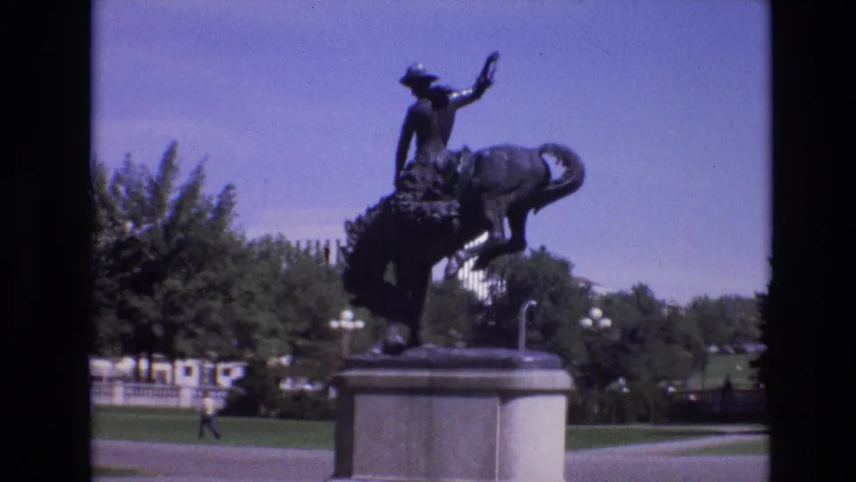 The home-movie camera continues on the Denver park statue, showing the rider, bucking horse, pedestal, nearby greenery, and a distant person crossing the walkway.