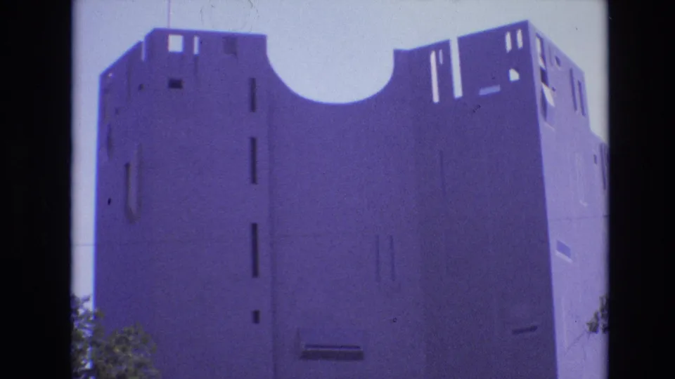 A 1974 Denver home-movie frame shows a pale modernist building facade with a large curved opening near the roofline, narrow rectangular cutouts, and trees at the lower edge.