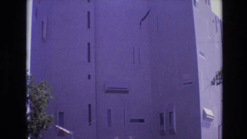 1974 Denver frame showing the upper portion of a stark concrete building, with slit windows, a pale sky, and a small tree partly visible below.