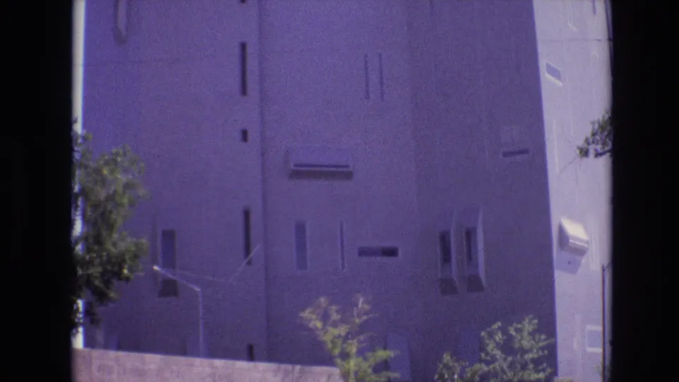 1974 Denver home-movie view of the lower and middle portions of the concrete building, with narrow windows, sparse trees, a streetlight, and heavy film grain.