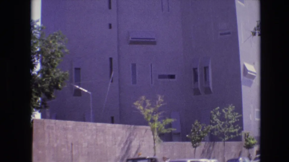 1974 Denver frame of the concrete tower from near its base, showing slit-like windows, a low wall, streetlight, young trees, and the purple cast of the 8mm scan.