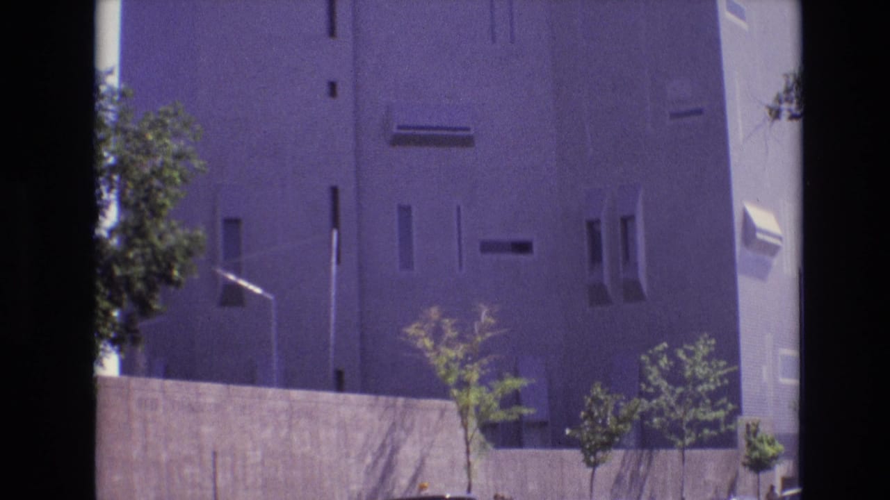 1974 Denver 8mm frame showing a tall modernist building facade, concrete wall, small trees, and a streetlight in bright daylight.