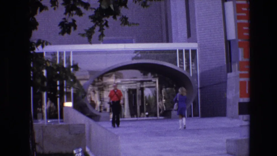 1974 Denver 8mm frame showing a man in a red shirt and a woman in a blue dress walking near an arched tunnel entrance and glass doors.
