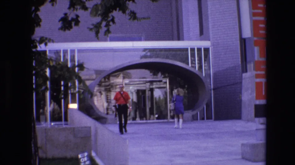 1974 Denver 8mm frame with two pedestrians crossing a concrete plaza-like sidewalk before a distinctive arched entrance and glass doorway.