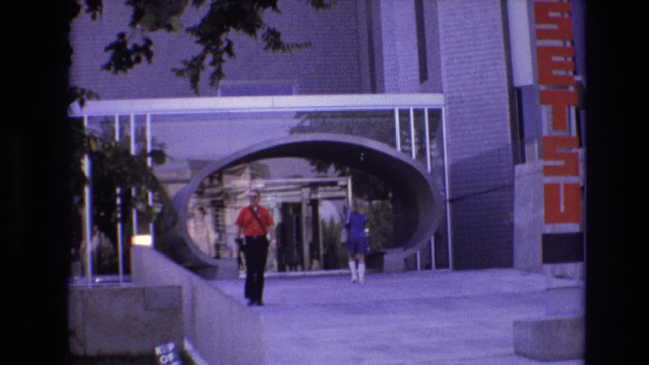 Man in a red shirt walks toward the camera near a modern arched building entrance.