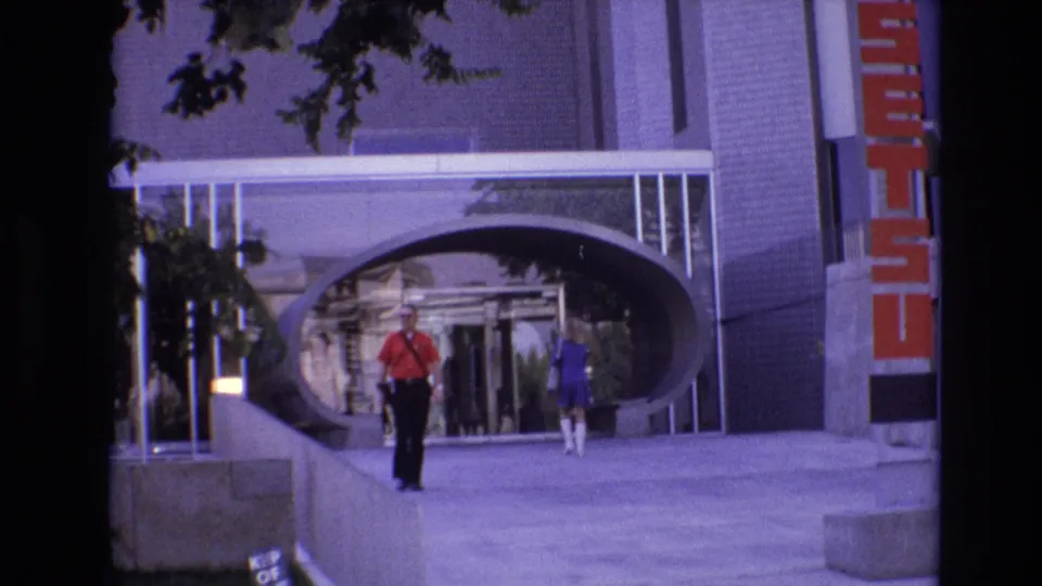 In 1974 Denver, a man in a red shirt walks along a sidewalk toward the camera while a woman in blue moves ahead near a brick-and-glass building entrance.
