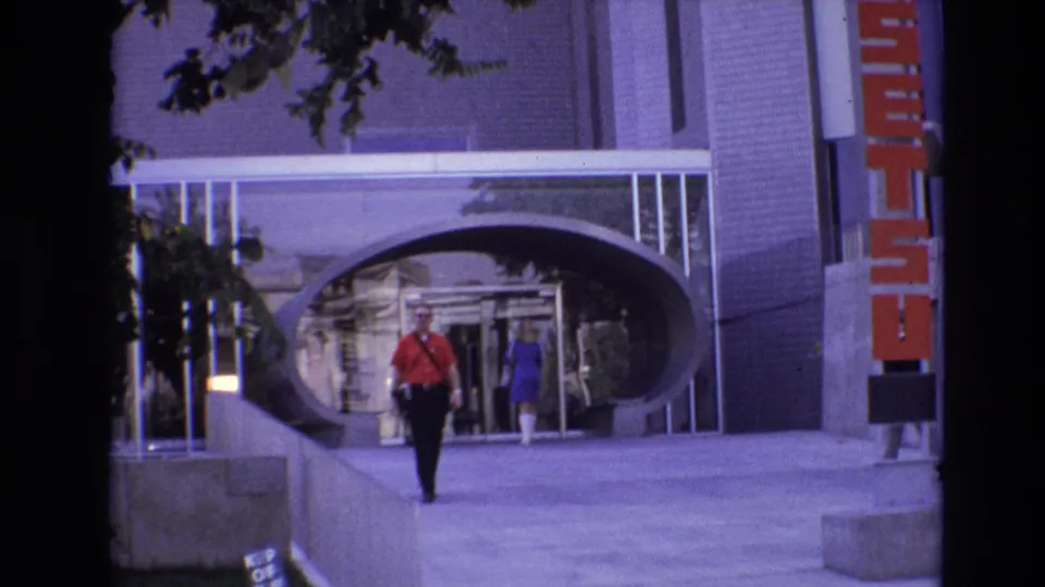 The Denver sidewalk view shows the man in red closer to the camera, with a woman in blue and reflective glass behind him under the oval-like entry arch.