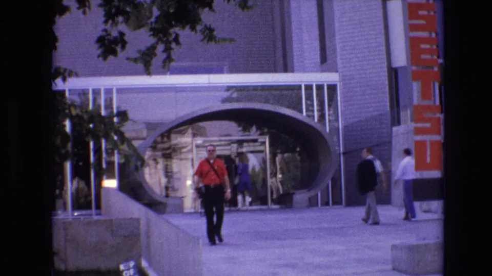 A closer 1974 Denver frame shows the red-shirted man walking toward the camera, with pedestrians in the background and the modern arched facade filling the scene.