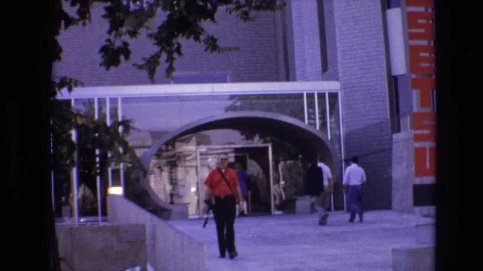 At the end of the Denver sidewalk clip, the man in red is near the foreground while two people pass to the right of the modern arched building entrance.
