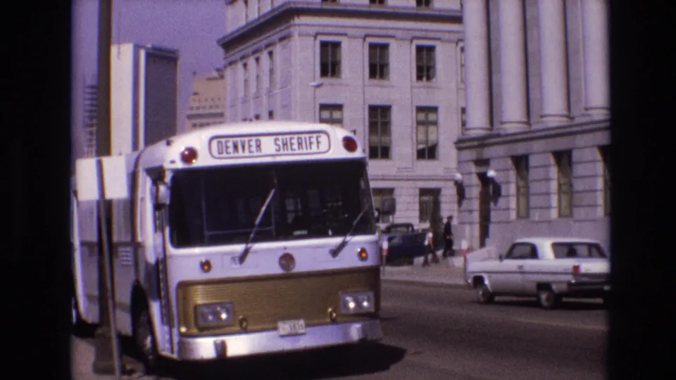 A white and gold Denver Sheriff bus is parked on a Denver city street in 1974, with large columns, pedestrians, and an older car nearby.