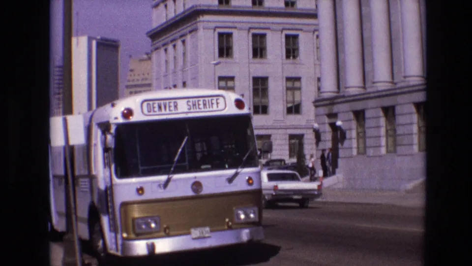 The front of the Denver Sheriff bus fills the frame on a 1974 Denver street, with civic-style architecture, pedestrians, and an older white car in the background.