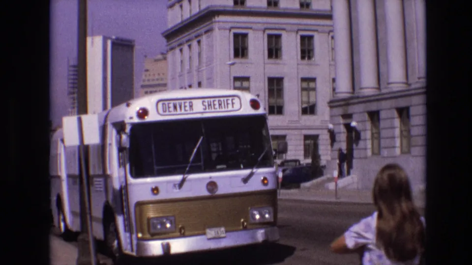 A 1974 Denver street frame shows the Denver Sheriff bus beside columned buildings, with a long-haired person seen from behind in the foreground.