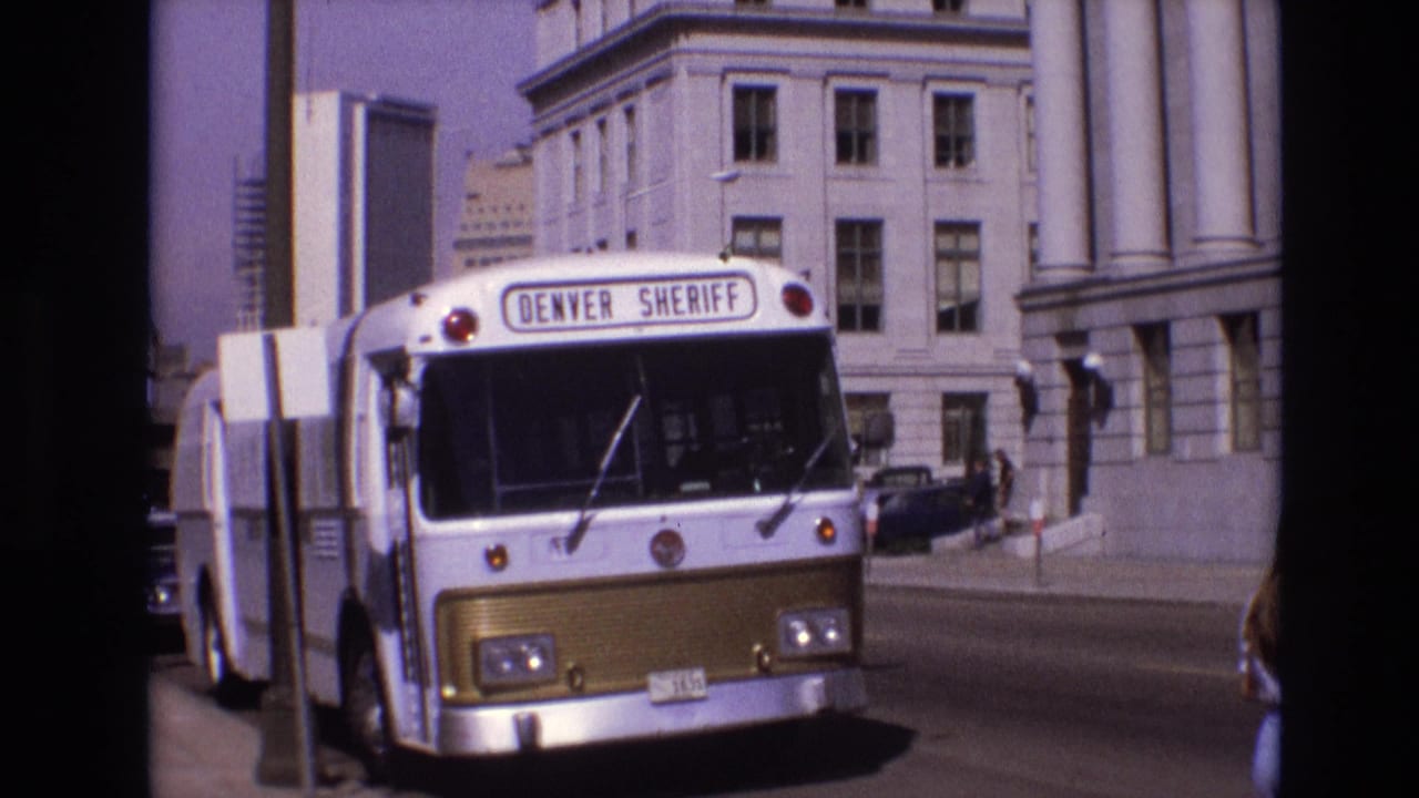 1974 Denver street view showing a white-and-gold Denver Sheriff bus near large columned buildings, with classic cars and sidewalk details in bright daylight.