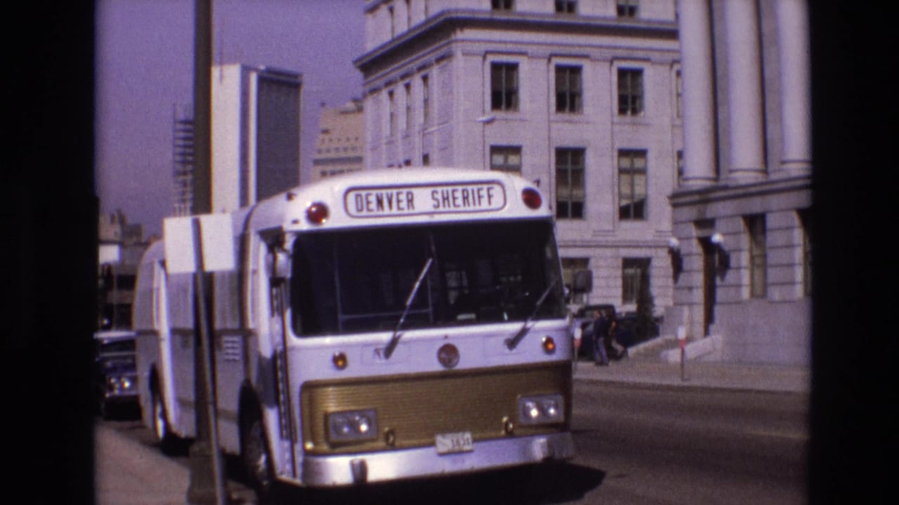 The Denver Sheriff bus remains parked or moving slowly near a columned building, with a visible pedestrian and period cars along the street in Denver.