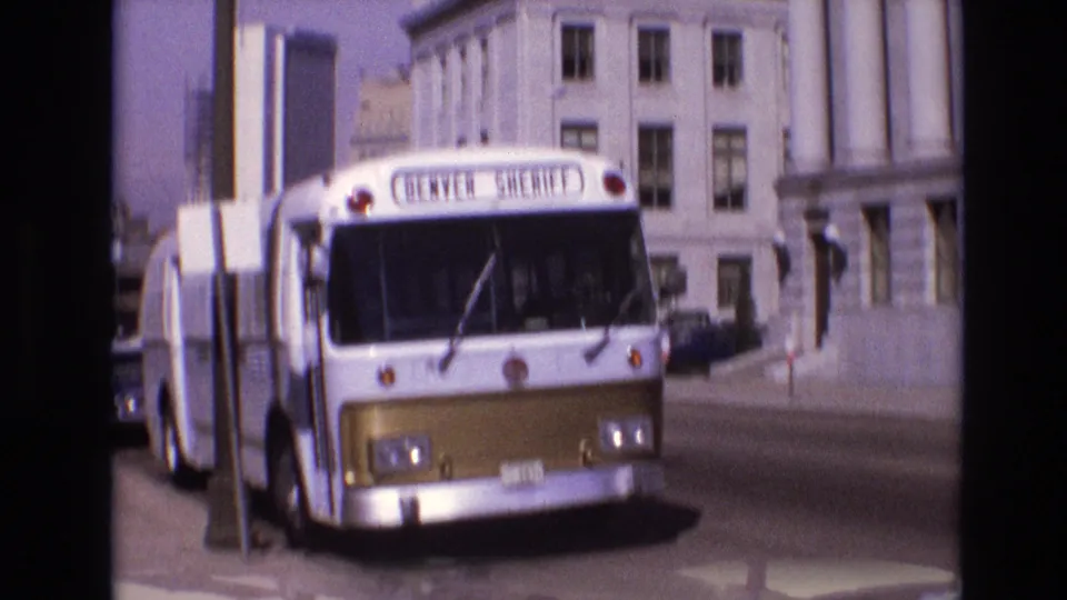 A softer 8mm frame shows the Denver Sheriff bus close to the curb, with civic-style architecture, street hardware, and traffic partially visible around it.
