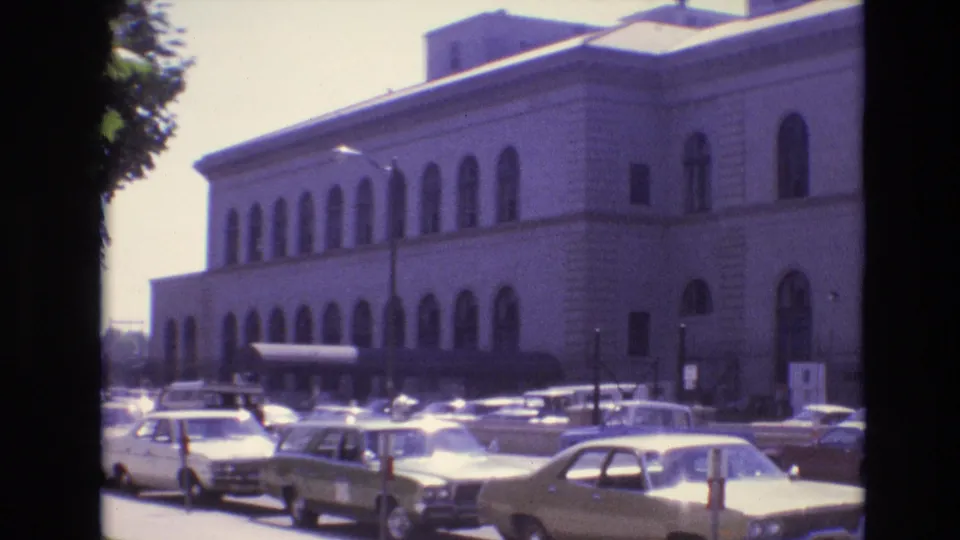 A 1974 Denver street-side view shows a crowded lot of classic cars in front of a large building with repeated arched windows and a shaded sidewalk area.