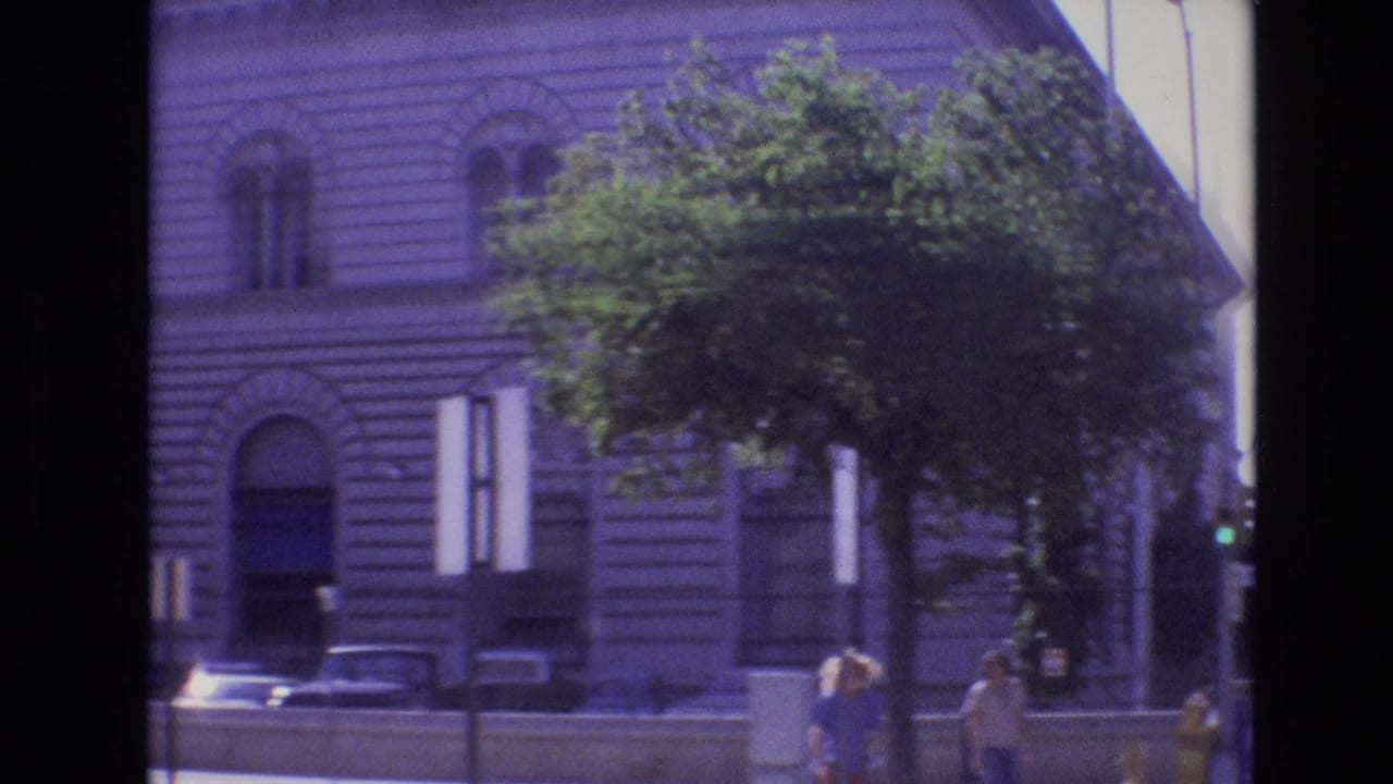 Large stone building with arched windows, trees, parked cars, and pedestrians along a street.
