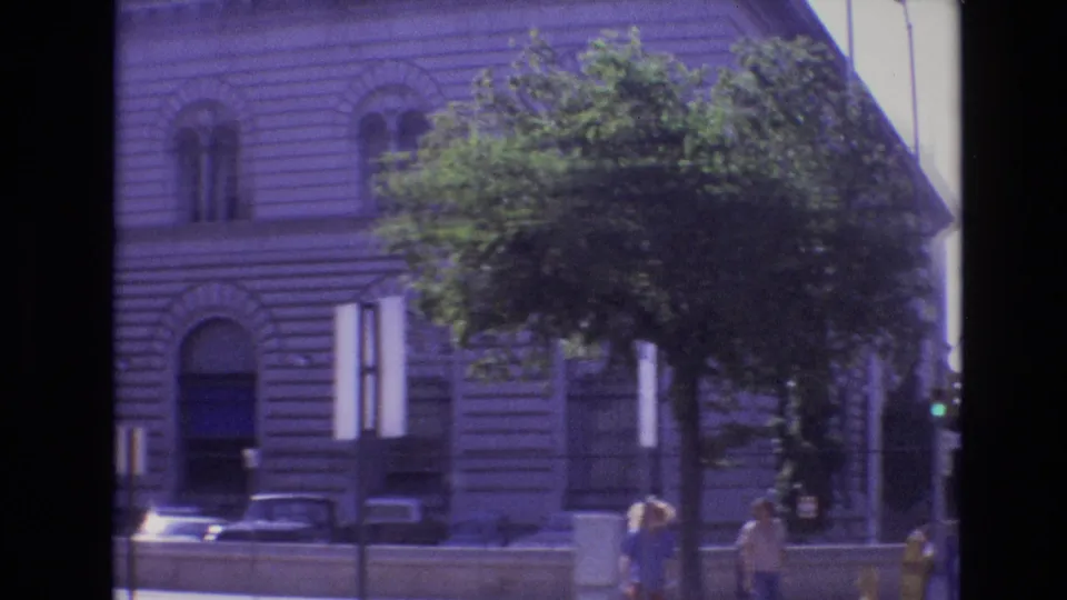 1974 Denver home-movie frame showing a large ornate stone building behind street trees, parked cars, and a few pedestrians on the sidewalk.