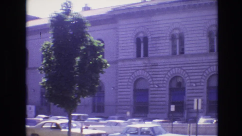1974 Denver home-movie frame with a street tree partly obscuring a large stone building, while classic cars sit parked along the curb.