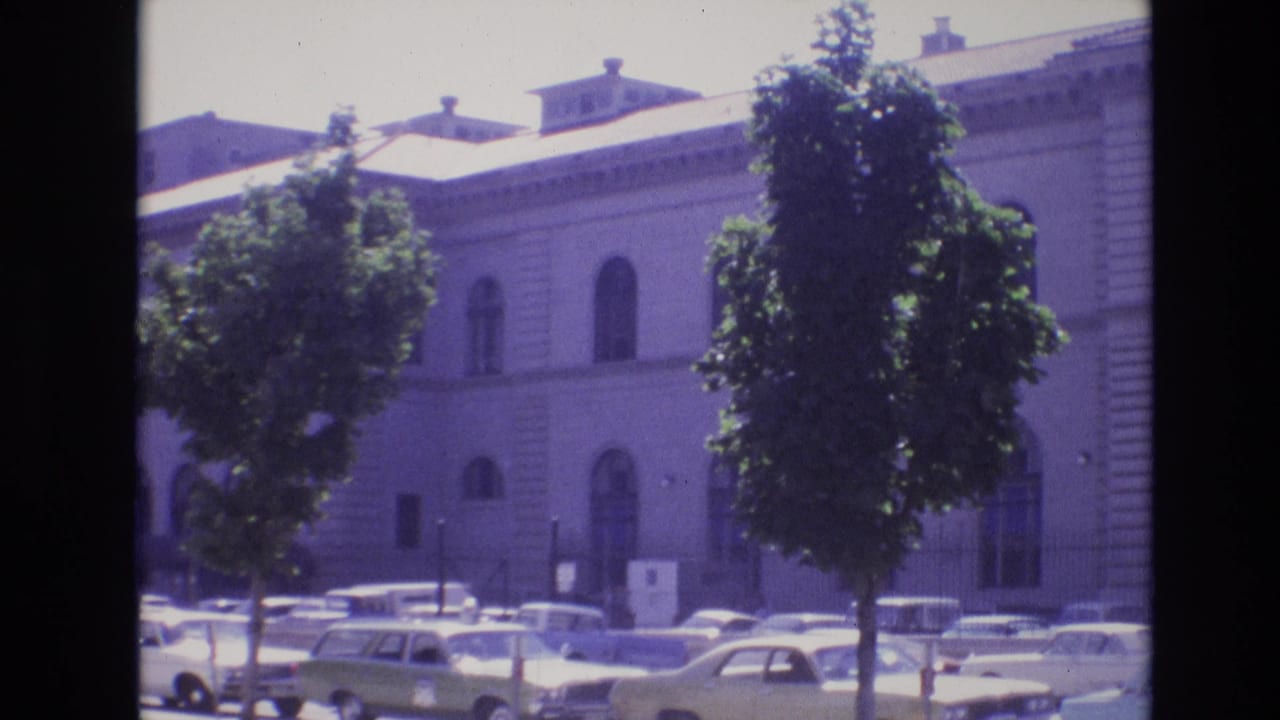 Large pale building with arched windows behind trees and parked cars.