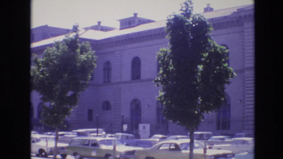 Denver 1974 home-movie frame showing a large pale institutional-looking building with arched windows, foreground trees, and parked 1970s cars.