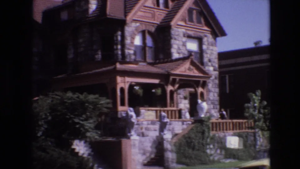 Denver 1974 home-movie frame showing an ornate stone-and-wood Victorian-style house exterior with a porch, front steps, shrubs, and decorative statues.