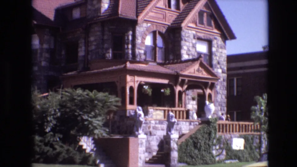 Denver 1974 exterior frame of a stone-and-wood house with porch railing, gabled upper windows, garden shrubs, decorative statues, and front steps.