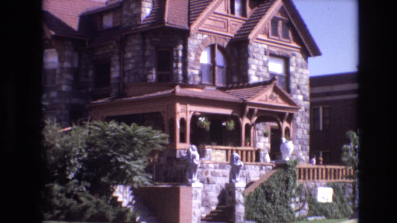 1974 Denver 8mm frame showing a large stone Victorian-style mansion with a brown wooden porch, arched window, white statues, and landscaped steps.