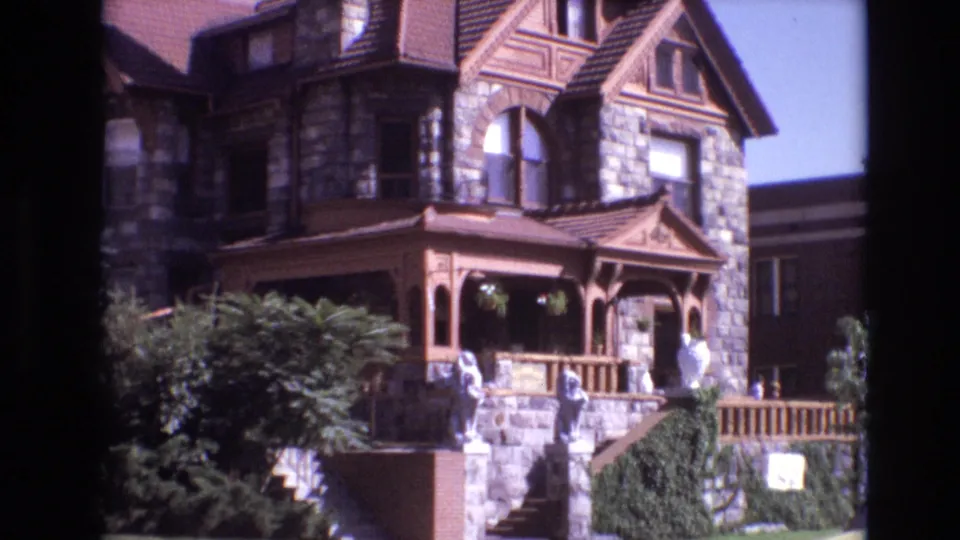 1974 Denver 8mm frame showing a large stone Victorian-style mansion with a brown wooden porch, arched window, white statues, and landscaped steps.