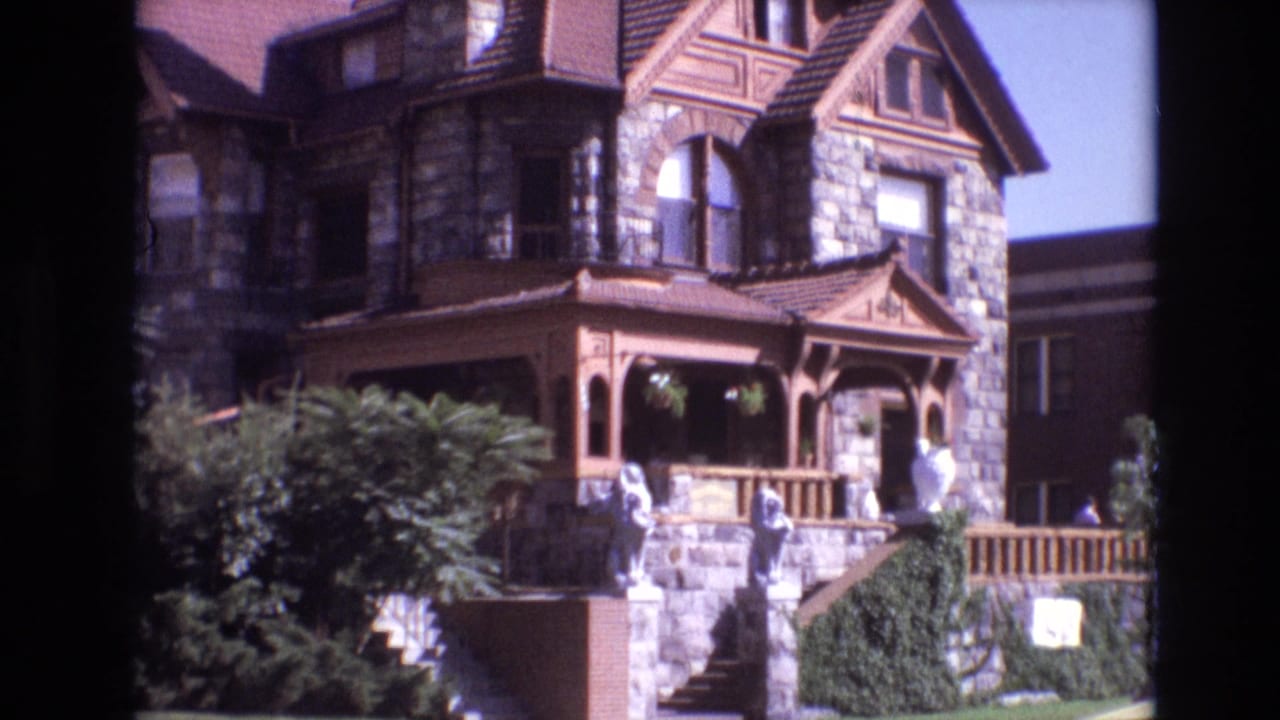 Stone mansion facade with ornate porch, statues on steps, shrubbery, and blue sky.