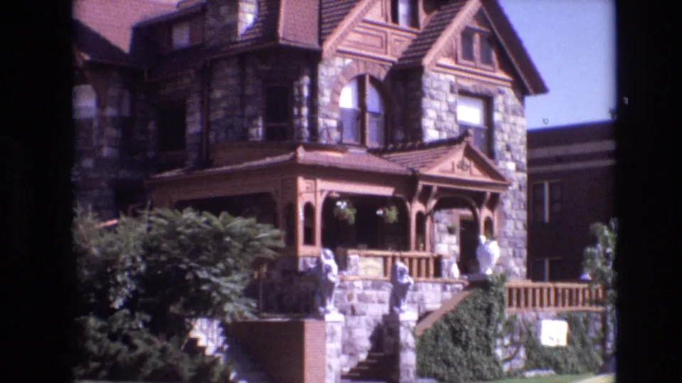 1974 Denver 8mm frame with a slightly closer view of the stone mansion's brown porch, steep gable, arched upper window, front steps, and surrounding greenery.