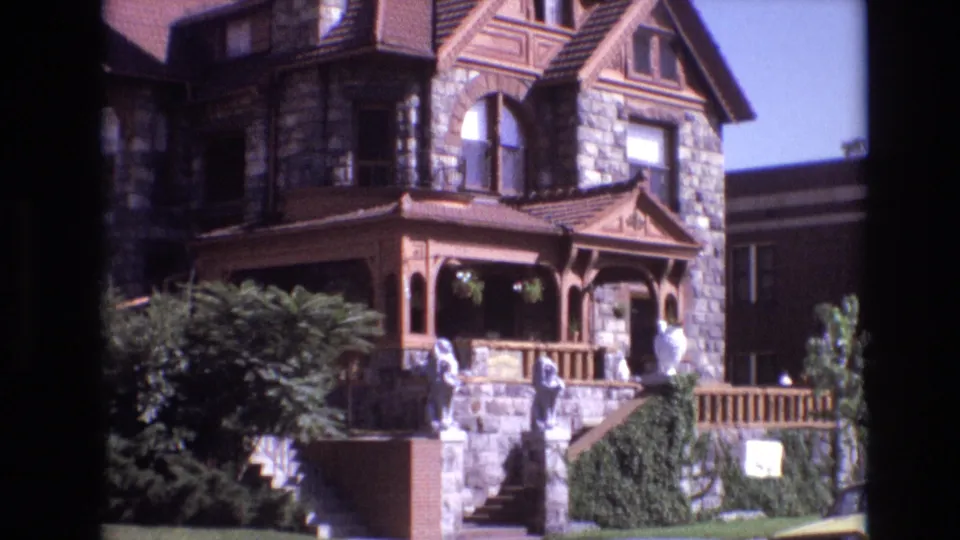Final 1974 Denver frame holding on the large stone mansion exterior, with the brown porch, white statues, front steps, clipped greenery, and adjacent structure visible.