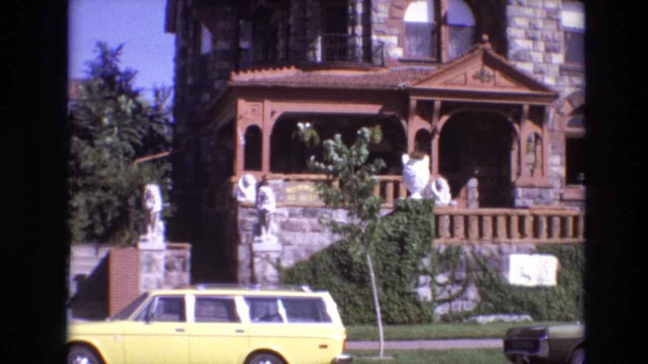 Stone building with wood porch, white statues, greenery, and a yellow station wagon parked along the street.