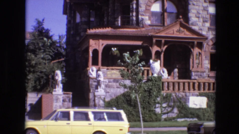 1974 Denver exterior view of a stone-and-wood building, porch statues, green landscaping, and a yellow station wagon passing across the lower frame.