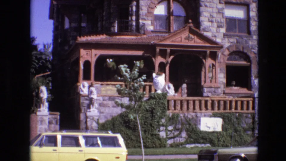 1974 Denver frame with a closer handheld view of the stone facade, decorative wood porch, white statues, leafy plants, and yellow station wagon along the street.