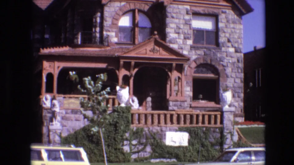 1974 Denver home-movie still showing the stone building and wood porch with statues and greenery, while a yellow station wagon and another car occupy the street foreground.