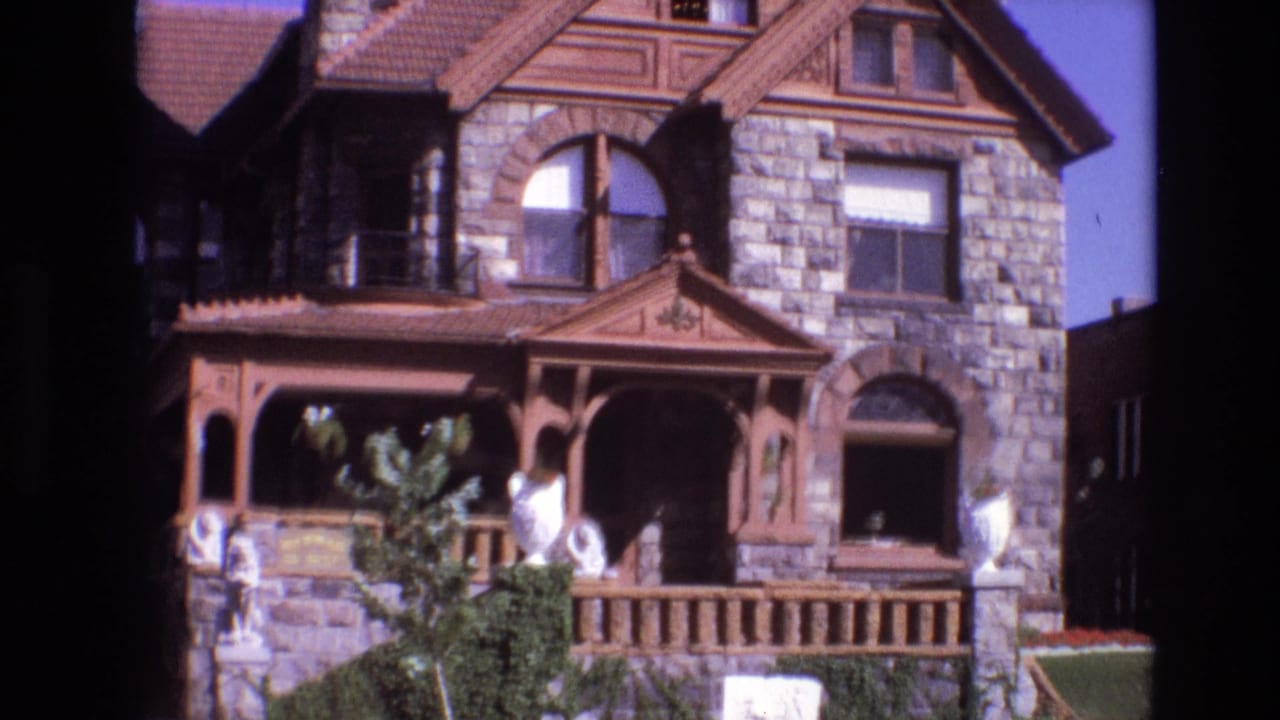 Stone Victorian-style house with wooden porch, arched windows, lawn, and people near the front entrance.