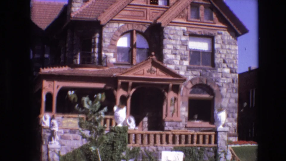 1974 Denver home-movie frame showing the front of a large stone Victorian-style house with a decorative wooden porch, arched windows, lawn, shrubs, and visible people near the entrance.