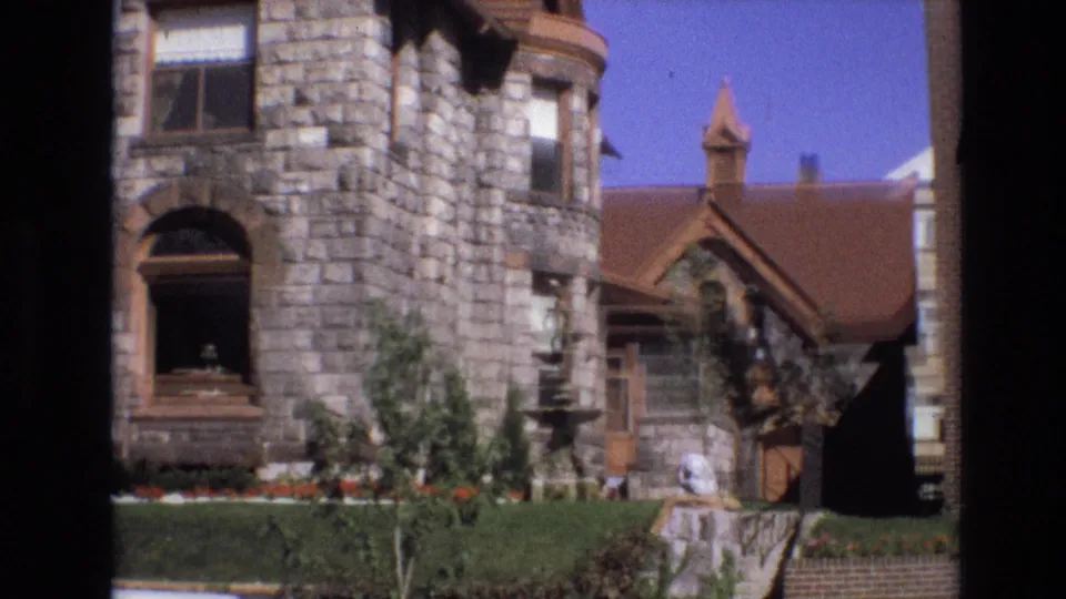 1974 Denver frame of the stone house from a slightly shifted angle, showing a turret-like corner, arched opening, fountain, clipped hedge, lawn, and side entry details.