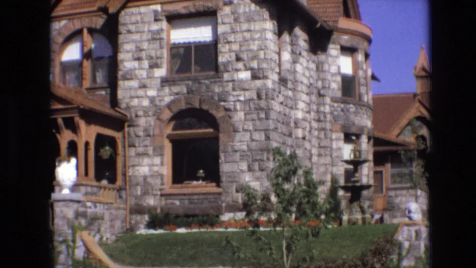 1974 Denver home-movie view of the stone house facade, including porch trim at left, arched and rectangular windows, a fountain, trimmed lawn, small trees, and planted beds.
