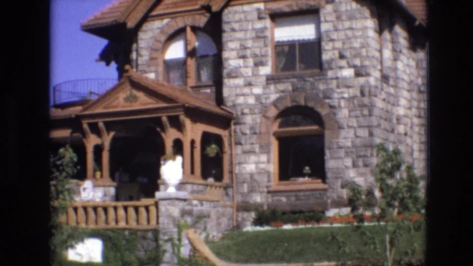 1974 Denver frame showing the front section of the ornate stone house with a decorative wooden porch, arched window, clipped hedge, lawn, small tree, roofline, and white statuary.