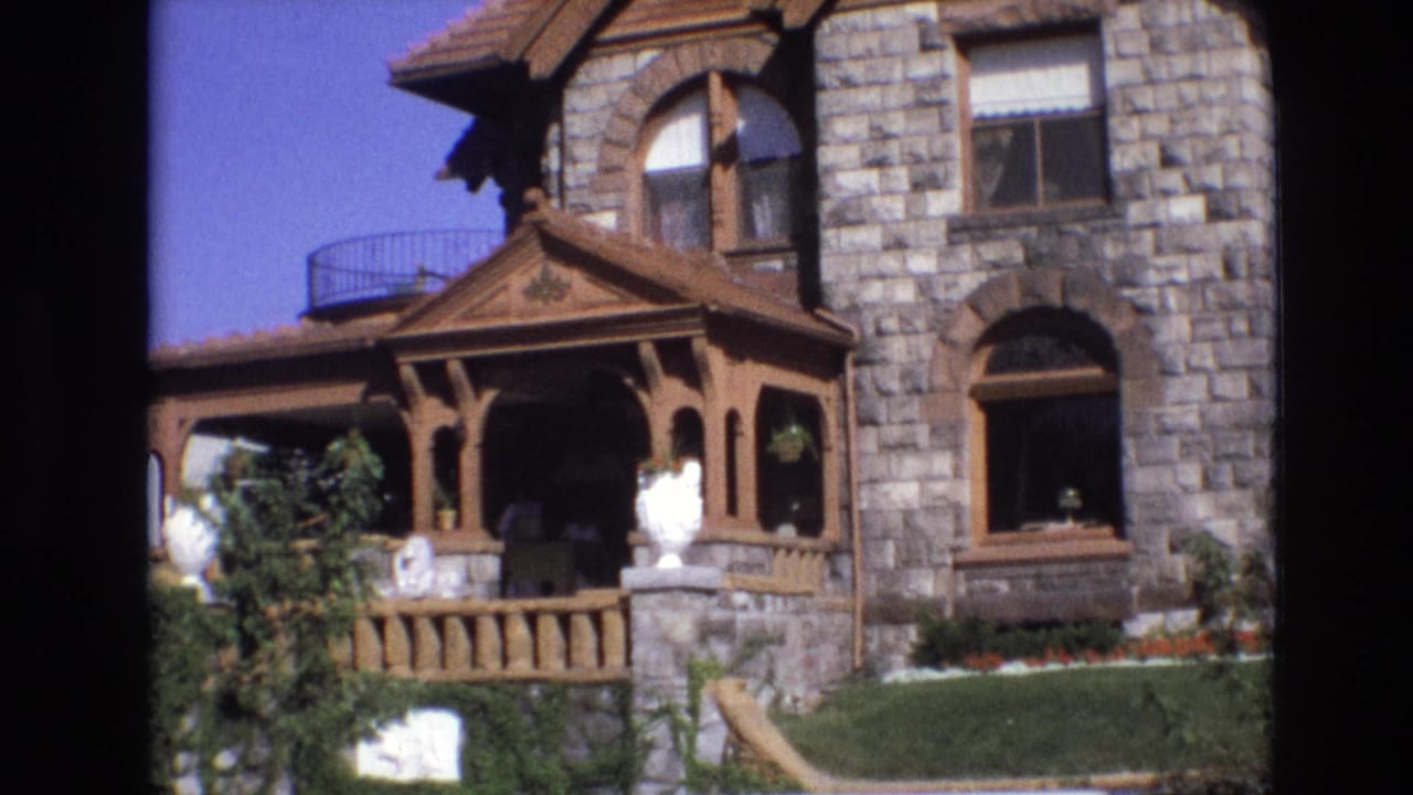 1974 Denver home-movie frame showing a stone-and-wood Victorian-style house exterior with a raised porch, arched windows, green landscaping, and white decorative urns.