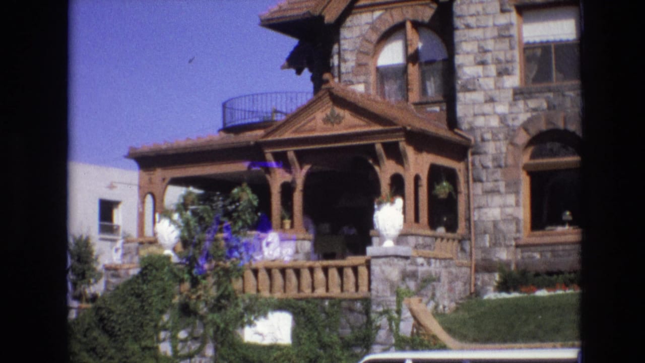 Porch and stone house facade with shrubs, white urns, adjacent building, and partial car roof.